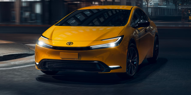 Yellow Toyota Prius driving on a city street at night with illuminated LED headlights